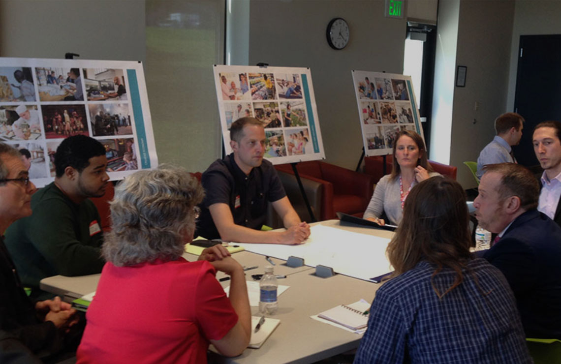 Community engagement workshop with architects and local stakeholders seated around a table, discussing plans and reviewing visual boards during a collaborative meeting.