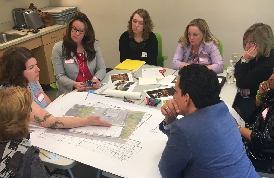 Conceptual design workshop with architects and stakeholders gathered around a table, reviewing drawings, site plans, and imagery while discussing early design ideas collaboratively.